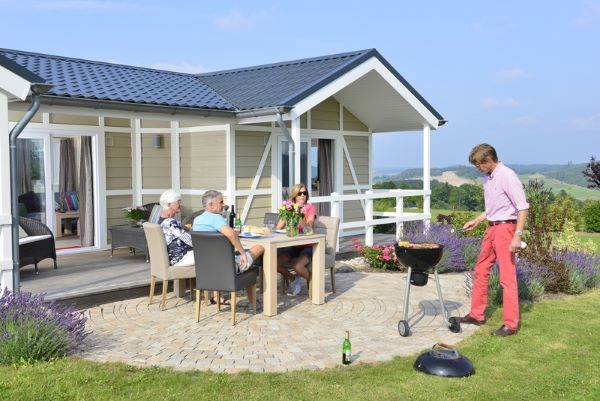 Eine Familie sitzt vor dem Chalet im Sauerland am Tisch und eine Person steht am Grill. Im Hintergrund ist eine weite Aussicht über das Sauerland sichtbar.